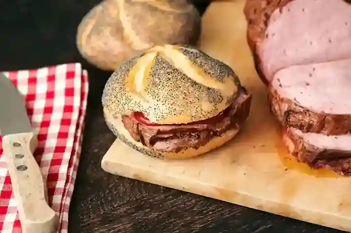 Frisches Mohnbrötchen mit hausgemachtem Leberkäse Backen, belegt mit Senf und Paprika, serviert auf einem Holzbrett neben frisch geschnittenem Leberkäse.