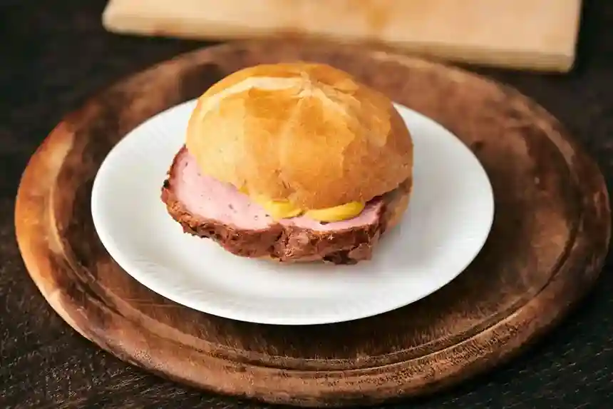 Frisches Brötchen mit einer dicken Scheibe Leberkäse Backen, serviert mit Senf auf einem weißen Teller und rustikalem Holzbrett.