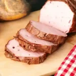 Frisch gebackener Leberkäse, in Scheiben geschnitten, auf einem Holzbrett serviert, mit knusprigen Brötchen und einem Messer – perfektes Leberkäse Backen Ergebnis.