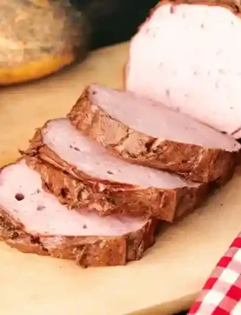 Frisch gebackener Leberkäse, in Scheiben geschnitten, auf einem Holzbrett serviert, mit knusprigen Brötchen und einem Messer – perfektes Leberkäse Backen Ergebnis.