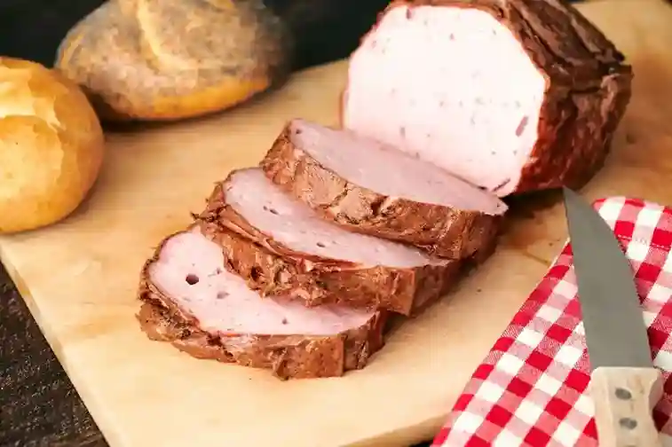 Frisch gebackener Leberkäse, in Scheiben geschnitten, auf einem Holzbrett serviert, mit knusprigen Brötchen und einem Messer – perfektes Leberkäse Backen Ergebnis.