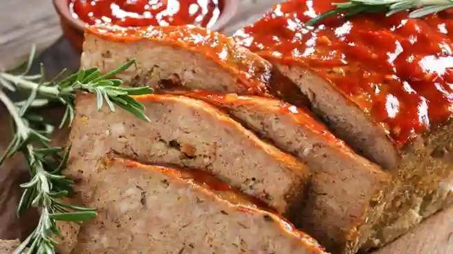 Gebackener Fleischkäse, in saftige Scheiben geschnitten, mit roter Glasur und frischem Rosmarin, angerichtet auf einem Holzbrett.