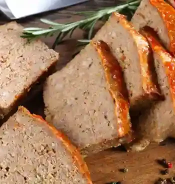 Gebackener Fleischkäse, in Scheiben geschnitten, mit einer knusprigen Kruste und roter Glasur, serviert auf einem Holzbrett, dekoriert mit Rosmarin.