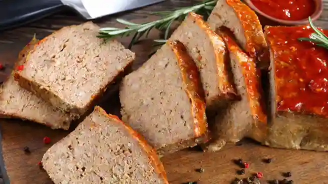 Gebackener Fleischkäse, in Scheiben geschnitten, mit einer knusprigen Kruste und roter Glasur, serviert auf einem Holzbrett, dekoriert mit Rosmarin.