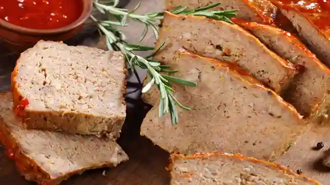 Gebackener Fleischkäse, in Scheiben geschnitten, mit glänzender roter Glasur und frischem Rosmarin, angerichtet auf einem Holzbrett.