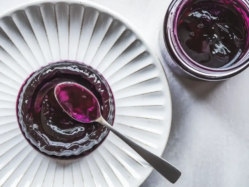 Zwetschgenmarmelade mit Lavendel auf einem Teller, mit einer Marmeladenflasche und einem Löffel daneben.
