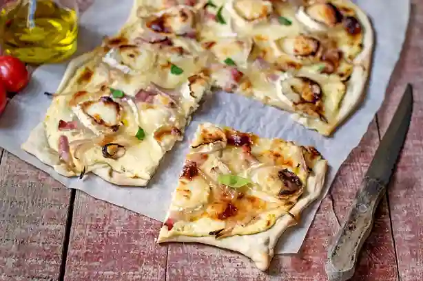 Knuspriger Flammkuchen vegetarisch mit Ziegenkäse, karamellisierten Zwiebeln und einem frischen Stück servierfertig auf Backpapier.