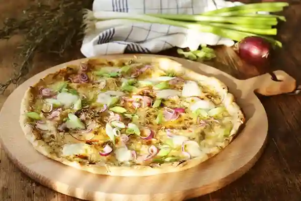 Frisch gebackener Flammkuchen vegetarisch auf einem runden Holzbrett, belegt mit roten Zwiebeln, Frühlingszwiebeln und Käse.