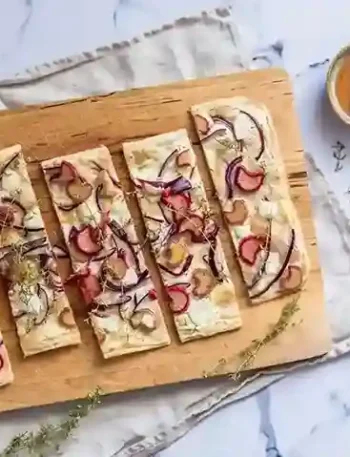 Flammkuchen mit Ziegenkäse, roten Zwiebeln, Birnen und Honig auf einem Holzbrett, daneben Honigtopf und Thymianzweige.