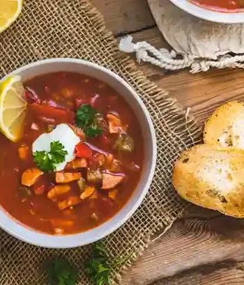 soljanka rezept oma ddr : Schüssel mit klassischer Soljanka nach Oma DDR Rezept, garniert mit saurer Sahne, Zitronenscheibe und frischer Petersilie, serviert mit knusprigem Brot auf rustikalem Holztisch.