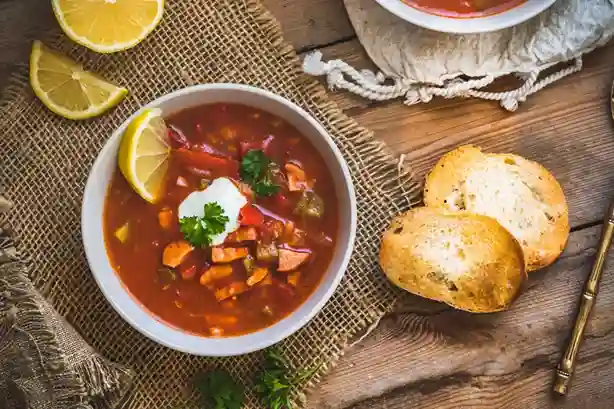 soljanka rezept oma ddr : Schüssel mit klassischer Soljanka nach Oma DDR Rezept, garniert mit saurer Sahne, Zitronenscheibe und frischer Petersilie, serviert mit knusprigem Brot auf rustikalem Holztisch.