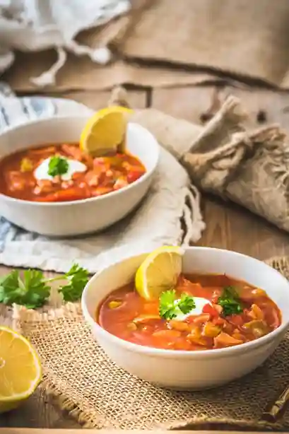 soljanka rezept oma ddr : Zwei Schüsseln mit klassischer Soljanka nach Oma DDR Rezept, garniert mit saurer Sahne, Zitronenscheiben und Petersilie, serviert auf rustikalem Holztisch mit Leinentuch.