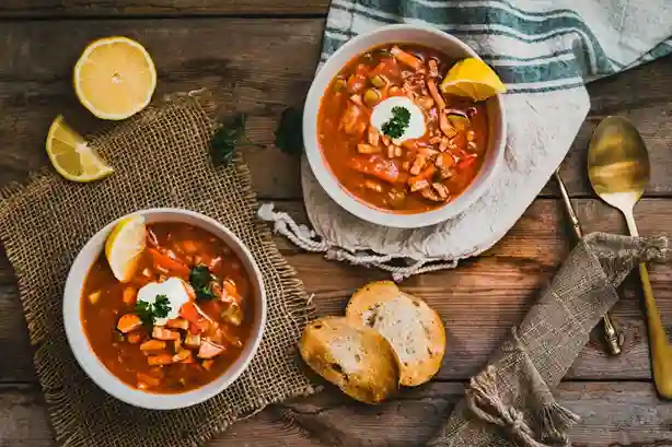 soljanka rezept oma ddr : Zwei Schüsseln mit traditioneller Soljanka nach Oma DDR Rezept, garniert mit saurer Sahne, Zitrone und Petersilie, serviert mit knusprigem Brot auf rustikalem Holztisch.