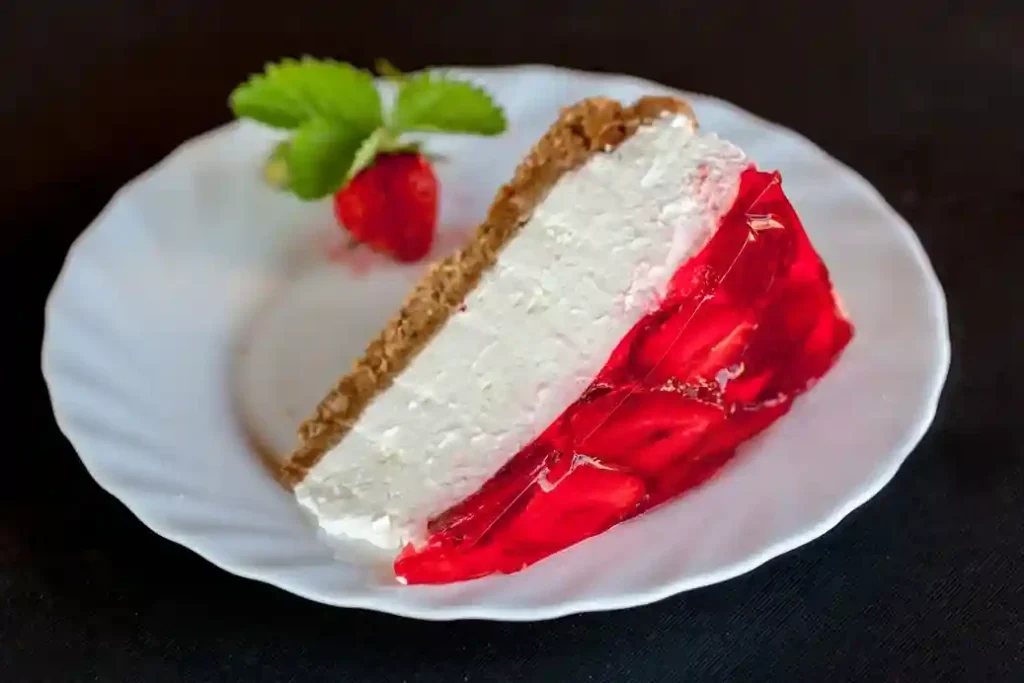 Ein Stück Erdbeerkuchen ohne Backen mit Keksboden und Mascarpone auf einem weißen Teller, mit einer frischen Erdbeere und Minzblättern dekoriert.