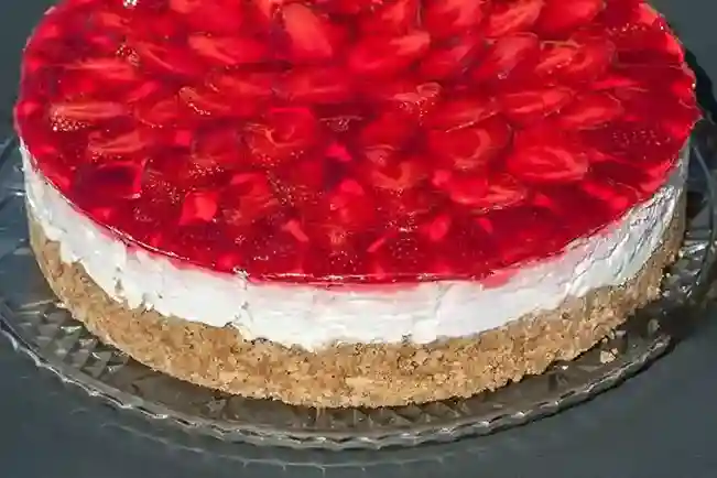 Ein klassischer Erdbeerkuchen ohne Backen mit Keksboden und Mascarpone, mit frischen Erdbeeren und glänzendem roten Tortenguss, auf einer Glasplatte serviert.