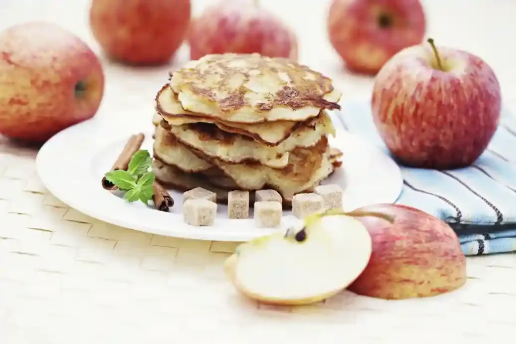 Ein Stapel frisch gebackener Apfelpfannkuchen auf einem Teller, umgeben von Äpfeln, Zimtstangen und Zuckerwürfeln – ein klassisches Apfelpfannkuchen Rezept.