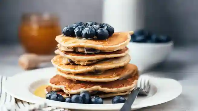 Ein Stapel Pfannkuchen vegan einfach, dekoriert mit Blaubeeren und Ahornsirup, serviert auf einem Teller, im Hintergrund ein Glas Honig und eine Schale Blaubeeren.