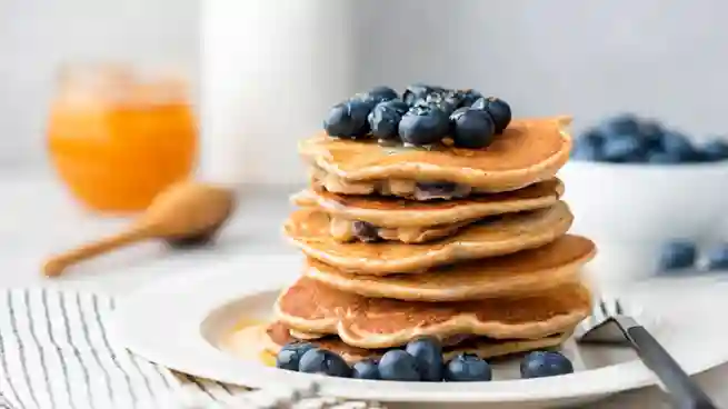 Ein Stapel Pfannkuchen vegan einfach, garniert mit Blaubeeren und Hanfsamen, serviert auf einem Teller, im Hintergrund ein Glas Honig und eine Schale Blaubeeren.