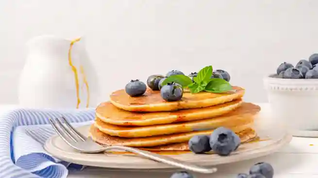 Ein Teller mit fluffigen Pfannkuchen vegan einfach, garniert mit frischen Blaubeeren und einem Minzzweig, daneben eine kleine Schale Blaubeeren und ein Krug.