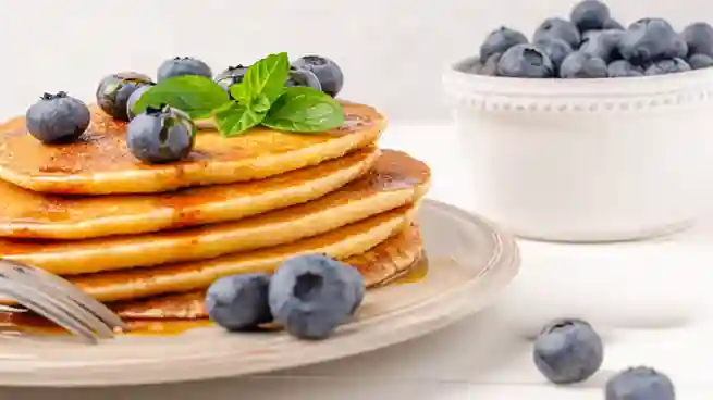 Ein Stapel fluffiger Pfannkuchen vegan einfach auf einem Teller, garniert mit frischen Blaubeeren und Minze, daneben eine weiße Schale voller Blaubeeren.