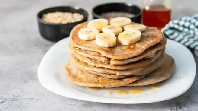 Ein Stapel Haferflocken Bananen Pancakes, garniert mit Bananenscheiben und Ahornsirup, auf einem weißen Teller.