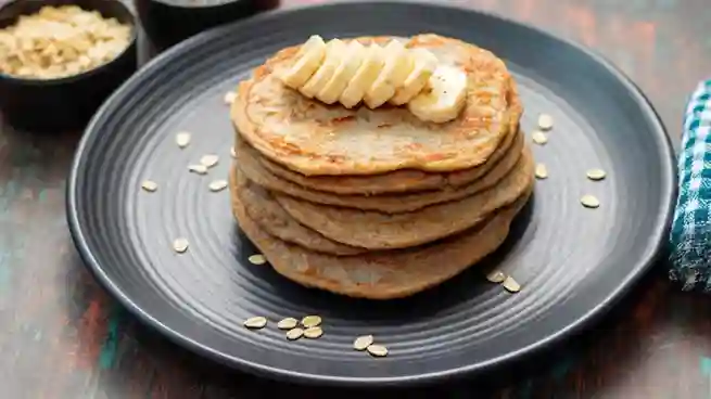 Ein Stapel Haferflocken Bananen Pancakes auf einem schwarzen Teller, garniert mit frischen Bananenscheiben und dekoriert mit Haferflocken.