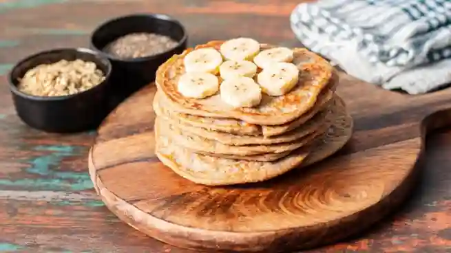 Ein Stapel Haferflocken Bananen Pancakes auf einem rustikalen Holzbrett, dekoriert mit Bananenscheiben, im Hintergrund Haferflocken und Chiasamen in Schalen.