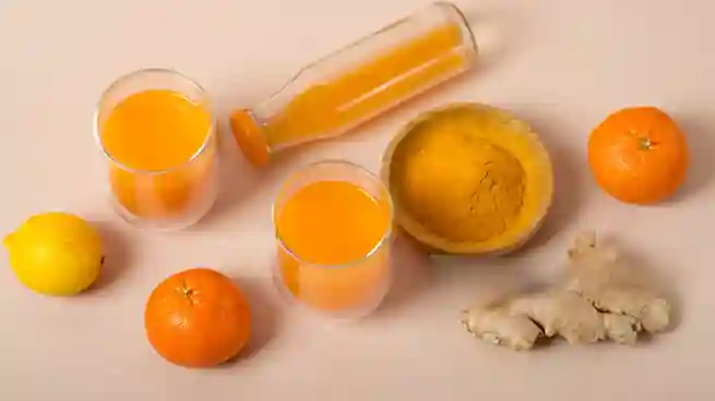 Zwei Gläser und eine Flasche selbstgemachter Ingwer Shot mit Orange und Kurkuma, umgeben von frischen Orangen, Zitronen, Ingwer und einer Schale Kurkumapulver.