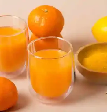 Zwei Gläser Ingwer Shot mit Orange und Kurkuma, umgeben von frischen Orangen, Zitronen, Ingwer und einer Schale mit Kurkumapulver.