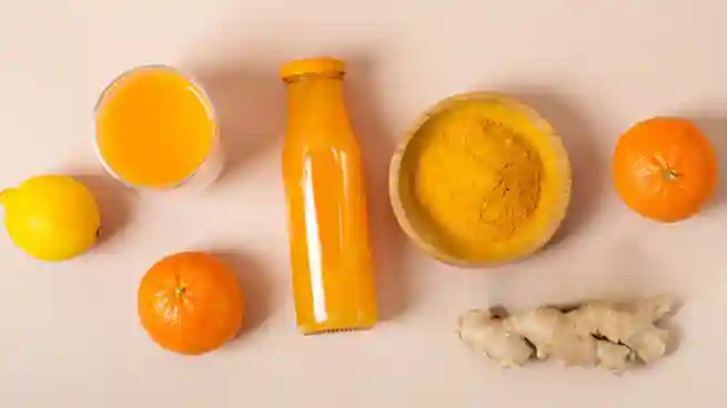 Eine Flasche und ein Glas selbstgemachter Ingwer Shot mit Orange und Kurkuma, umgeben von frischen Orangen, Zitronen, Ingwer und einer Schale mit Kurkumapulver.