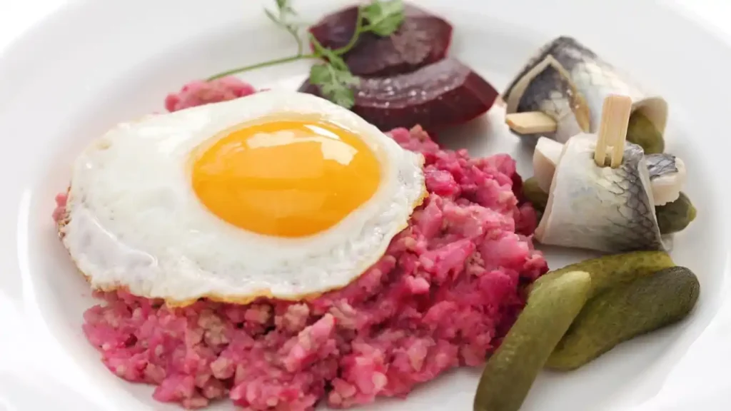 Labskaus Rezept serviert mit Spiegelei, Rote Bete, Rollmops und Gewürzgurken auf einem weißen Teller.