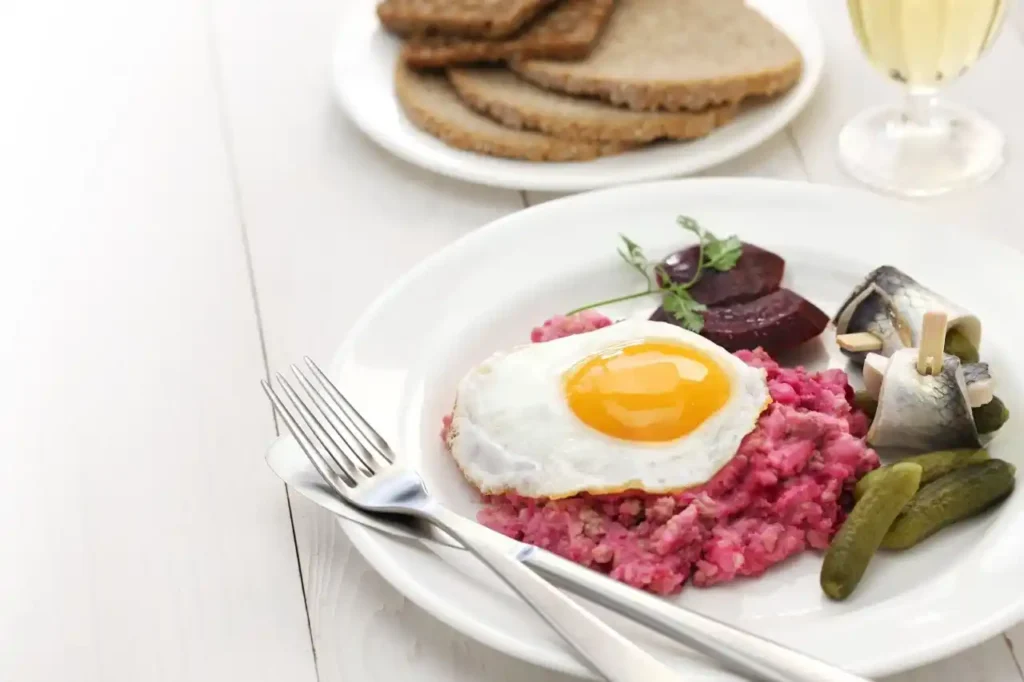 Labskaus Rezept serviert mit Spiegelei, Rote Bete, Rollmops und Gewürzgurken auf einem weißen Teller.