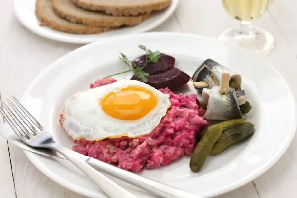 Labskaus Rezept – Traditionelles norddeutsches Gericht mit Spiegelei, Gewürzgurken, Roter Bete und Rollmops, serviert auf einem weißen Teller.