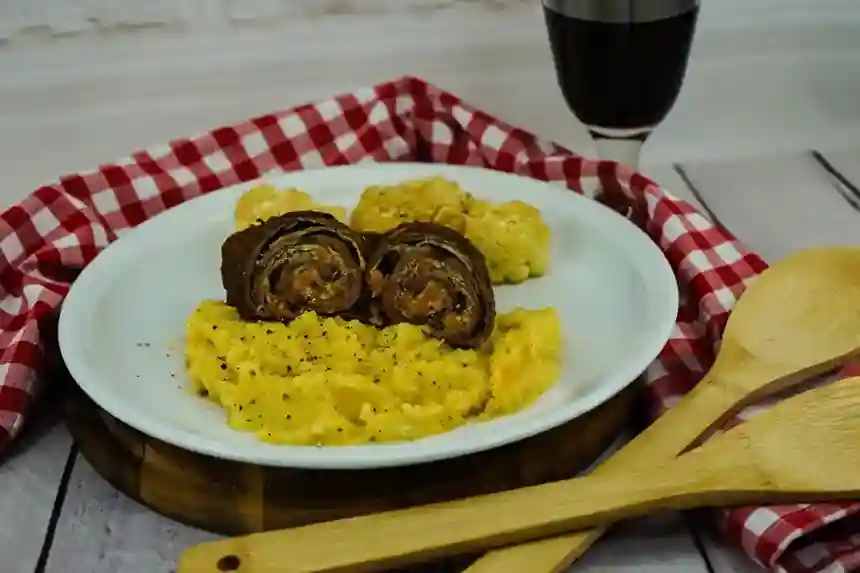 Aufgeschnittene Rouladen im Schnellkochtopf serviert mit cremigem Kartoffelpüree, geröstetem Blumenkohl und einem Glas Rotwein auf einem rustikalen Holztisch.