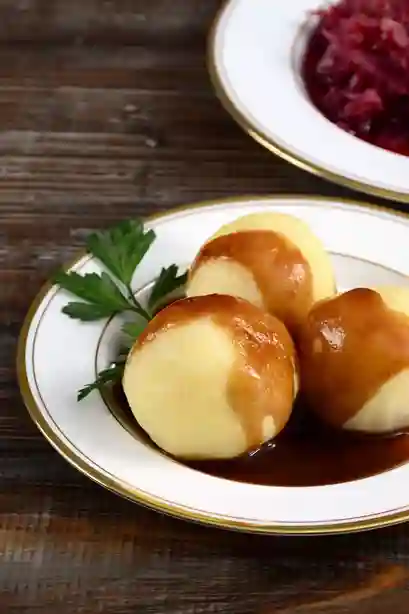 Drei Kartoffelklöße mit dunkler Sauce für Knödel, serviert auf einem klassischen weißen Teller mit goldener Umrandung.