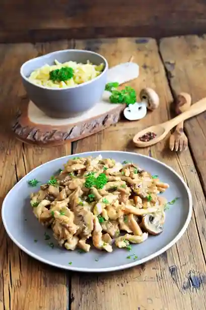 Spätzle mit Hähnchen in cremiger Pilzsoße, serviert mit einer Schüssel Spätzle auf einem rustikalen Holztisch.