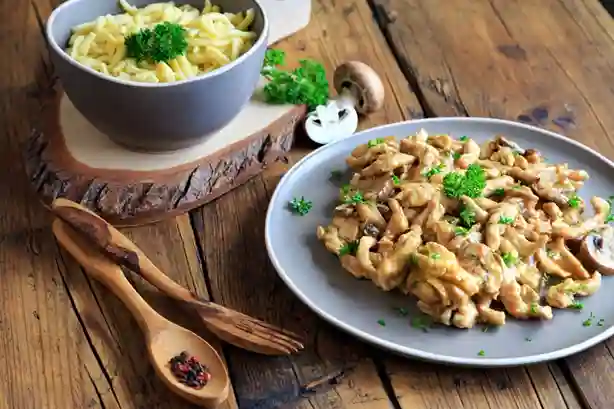 Spätzle mit Hähnchen und Champignons in cremiger Soße, serviert mit einer Schüssel Spätzle auf rustikalem Holztisch.