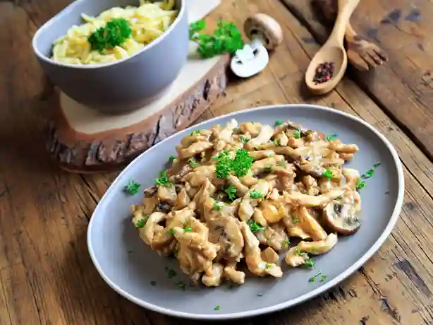 Spätzle mit Hähnchen und Pilzrahmsoße, serviert mit einer Schüssel Spätzle auf einem rustikalen Holztisch.