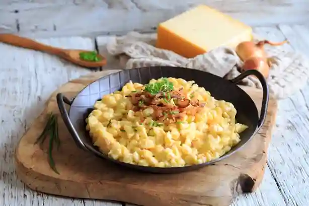 Schwäbische Käsespätzle Pfanne mit geschmolzenem Käse und knusprigen Röstzwiebeln, serviert in einer schwarzen Pfanne auf einem rustikalen Holzbrett.