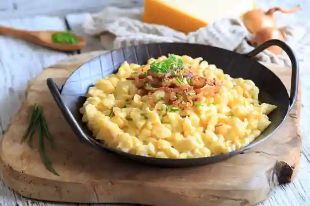 Schwäbische Käsespätzle Pfanne mit goldbraunen Röstzwiebeln, serviert in einer schwarzen Pfanne auf einem Holzbrett.