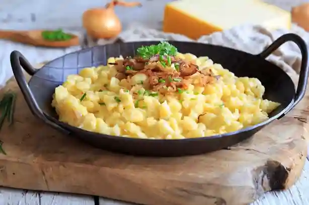 Schwäbische Käsespätzle Pfanne mit geschmolzenem Käse, knusprigen Röstzwiebeln und frischer Petersilie, serviert in einer schwarzen Pfanne auf einem rustikalen Holzbrett.