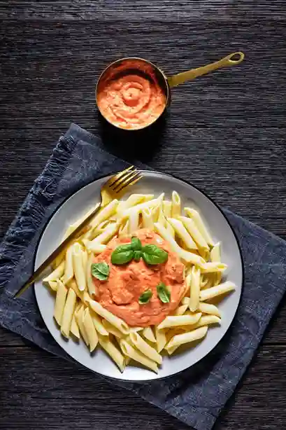 Penne mit Tomaten Sahne Soße und frischem Basilikum auf einem Teller, daneben ein Topf mit cremiger Sauce.