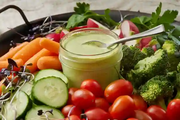 Frisches Avocado Dressing in einem Glas, serviert mit geschnittenem Gemüse wie Gurken, Karotten, Tomaten und Brokkoli.