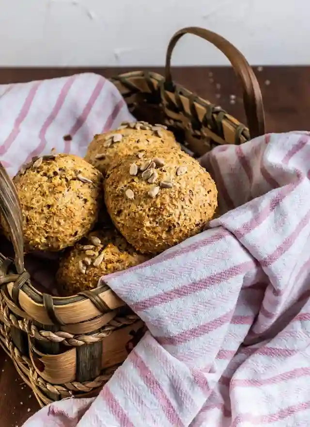 Selbst gebackene schnelle Sonntagsbrötchen ohne Hefe mit Körnern in einem rustikalen Brotkorb auf gestreiftem Küchentuch