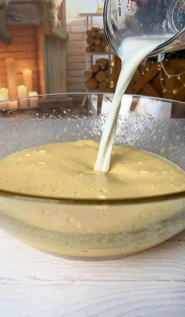Warme Milch wird in eine Glasschüssel mit schaumiger Eier-Zucker-Masse für französischer unsichtbarer Apfelkuchen gegossen.