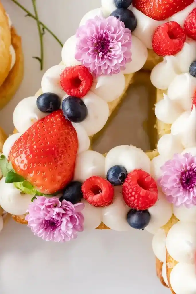 Nahaufnahme einer Zahlentorte mit Biskuitboden, dekoriert mit frischen Beeren, Sahnecreme und essbaren Blumen – Number Cake in Detailansicht