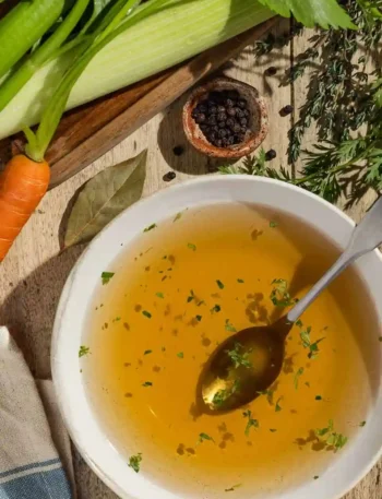 Hausgemachte Gemüsebrühe ohne Salz in einer Schüssel, umgeben von frischem Bio-Gemüse, Lorbeerblatt und Pfeffer – fettfrei, glutenfrei und perfekt zum Abnehmen