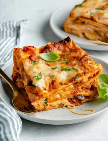 Vegane Lasagne Bolognese mit Gemüse und geschmolzenem veganem Käse auf einem weißen Teller, serviert mit frischem Basilikum