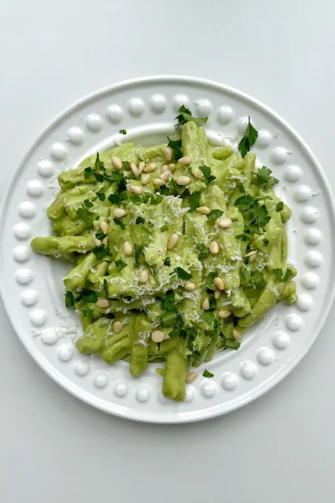 Pasta mit cremiger Avocado-Basilikum-Soße auf weißem Teller, garniert mit Parmesan, Pinienkernen und frischen Kräutern – eine moderne, leckere Soße für Pasta.