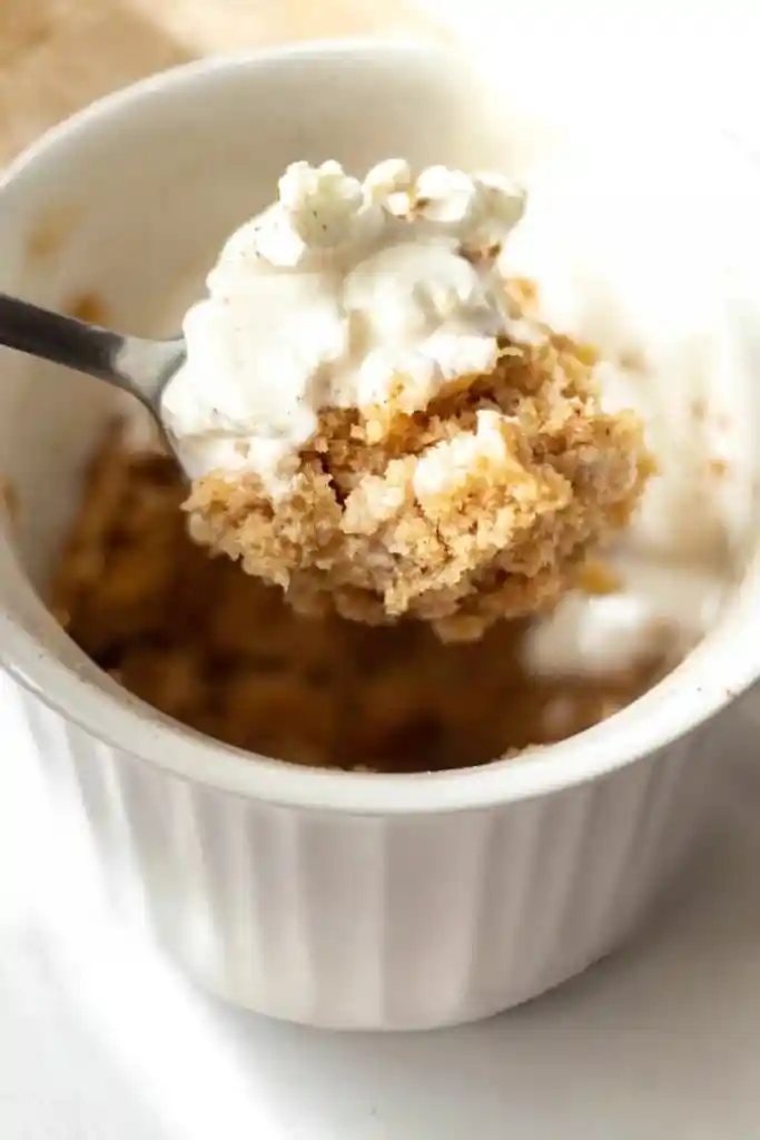 Löffel mit Apfel-Zimt-Tassenkuchen und Schlagsahne über einer weißen Keramiktasse – schneller süßer Snack mit wenigen Zutaten, in 5 Minuten zubereitet.